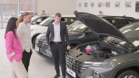 young car salesman showing to young couple new automobile at dealership salon.