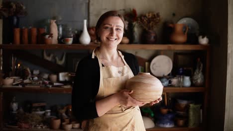 light brown vase from clay in the female potter's hands. portrait of cheerful blonde woman holding a vase of clay. the potter works in a pottery workshop with clay. front view