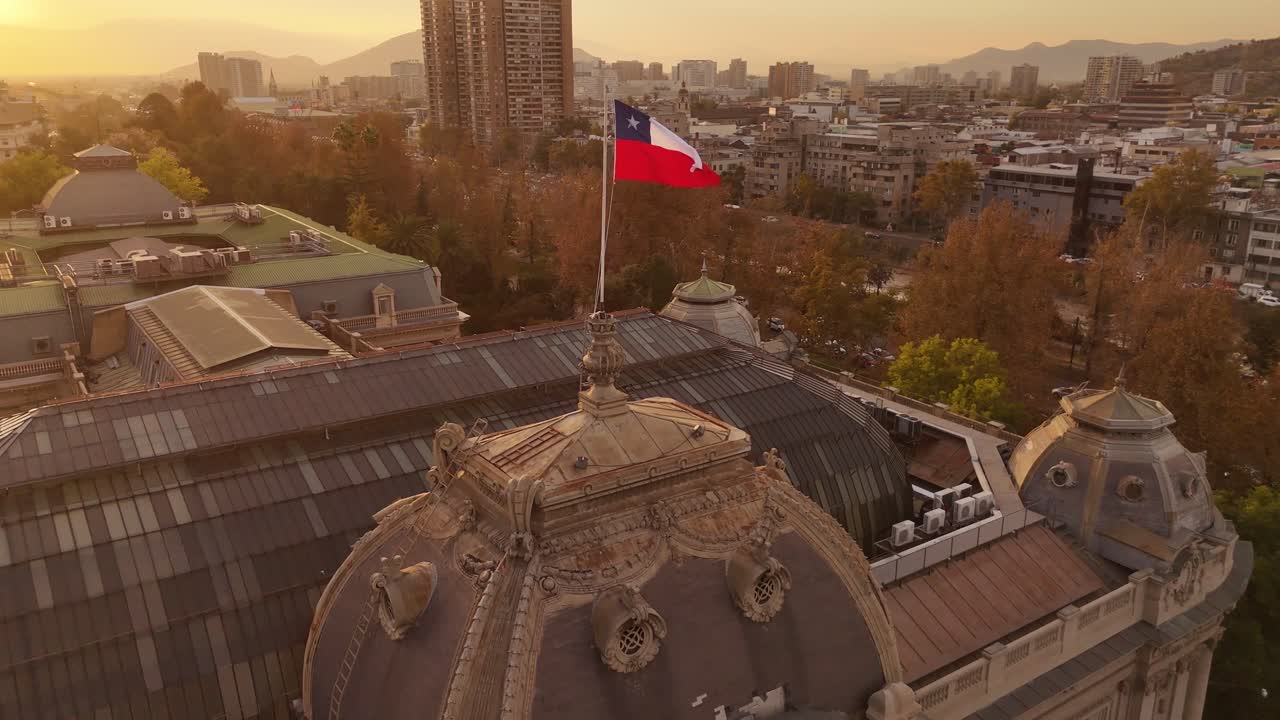 Aerial establishing shot of flying flag of Chile at golden sunset. de Chile City with tower and golden trees. Wide shot.