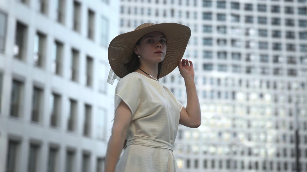 Una donna elegante è vista di spalle all'aperto in un ambiente urbano luminoso, che gira il viso sopra la spalla con alti edifici per uffici leggermente sfocati sullo sfondo