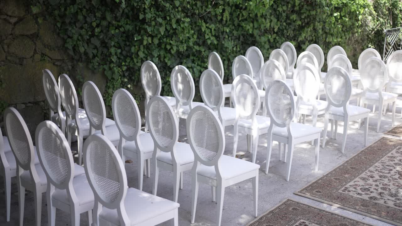 sillas blancas dispuestas para un evento al aire libre contra una pared de piedra cubierta de hiedra