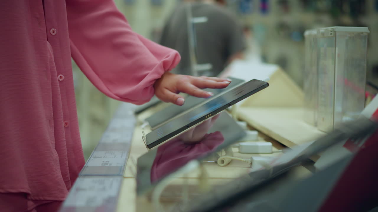 핑크색 드레스를 입은 손이 밝은 기술 상점의 디스플레이 스탠드에 있는 태블릿과 상호 작용하며, 화면이 작동되는 동안 장치가 끔한 스탠드에서 다른 사람의 흐릿한 시야로 앉아 있습니다.