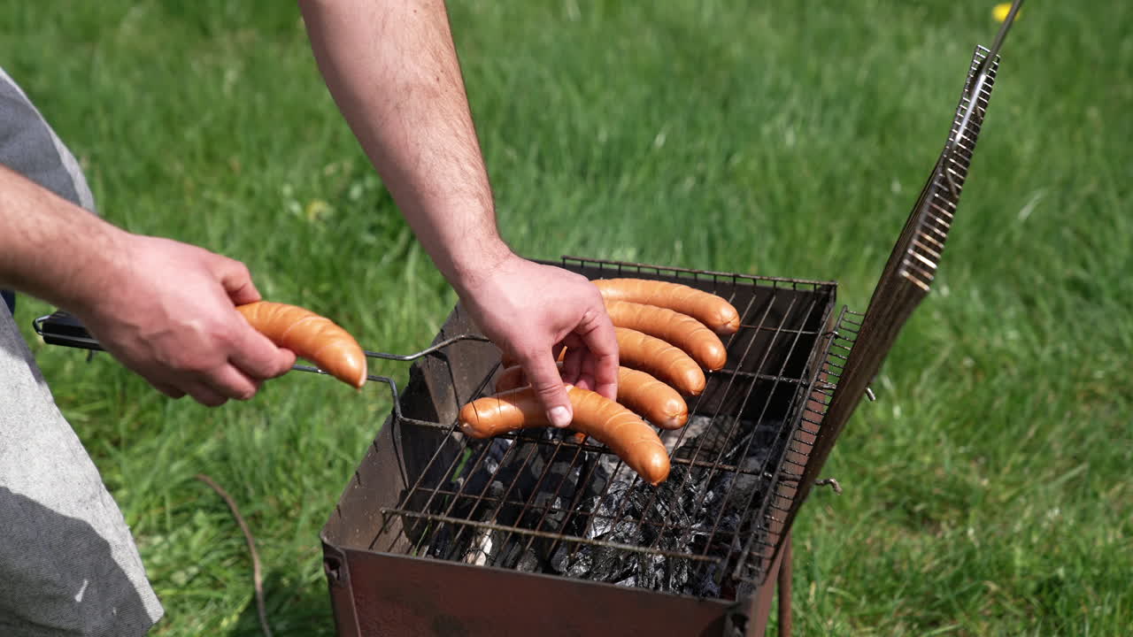 Sausages roasting on barbecue grill outdoors. Grilled food fried and smoked in charcoal grills. Man chef putting delicious sausages on a grill grate. Food for picnic.