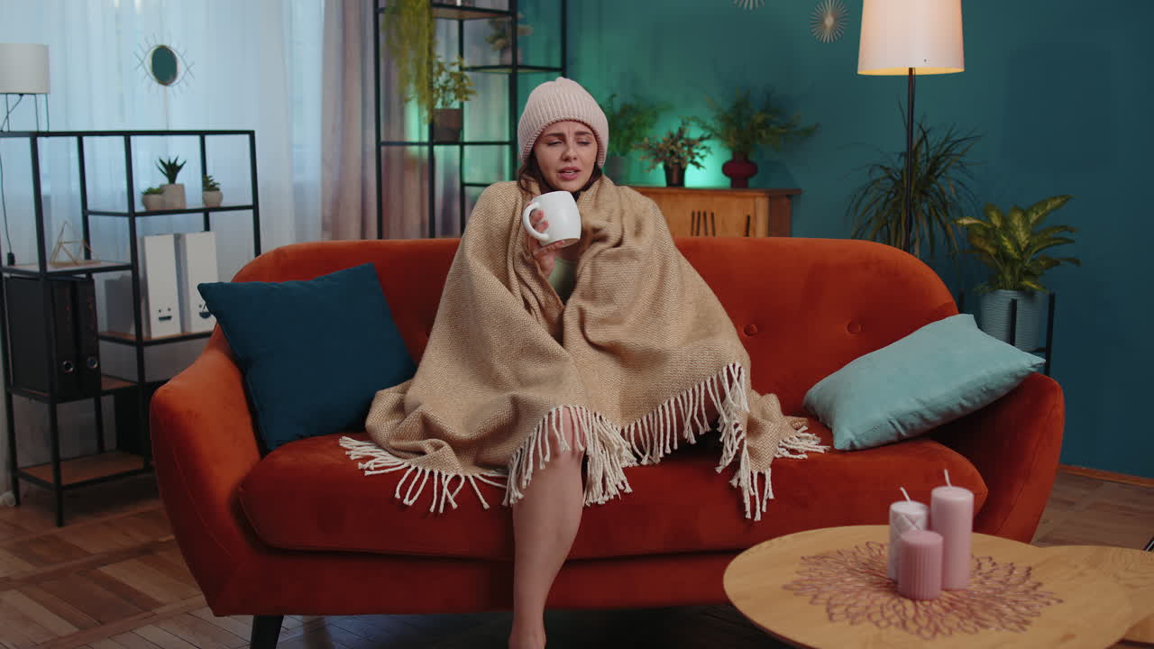 una joven enferma usa sombrero envuelto en cuadros siéntase sola temblando de frío en el sofá bebiendo té caliente