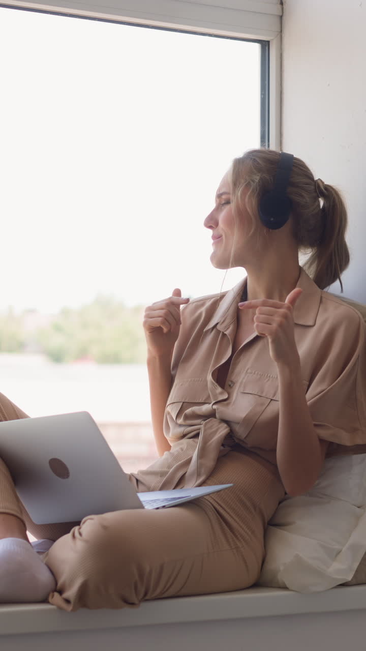 Young blonde woman in headphones has fun listening to pop dancing music sitting on windowsill leaned against cushions in comfortable room slow motion