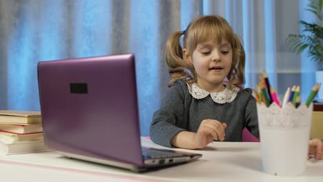 child girl doing school homework writes in notebook, using laptop computer at home, online education