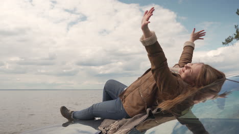 red haired woman lying on the bonnet of her car next to a beautiful lake, she covers the clouds with her hands