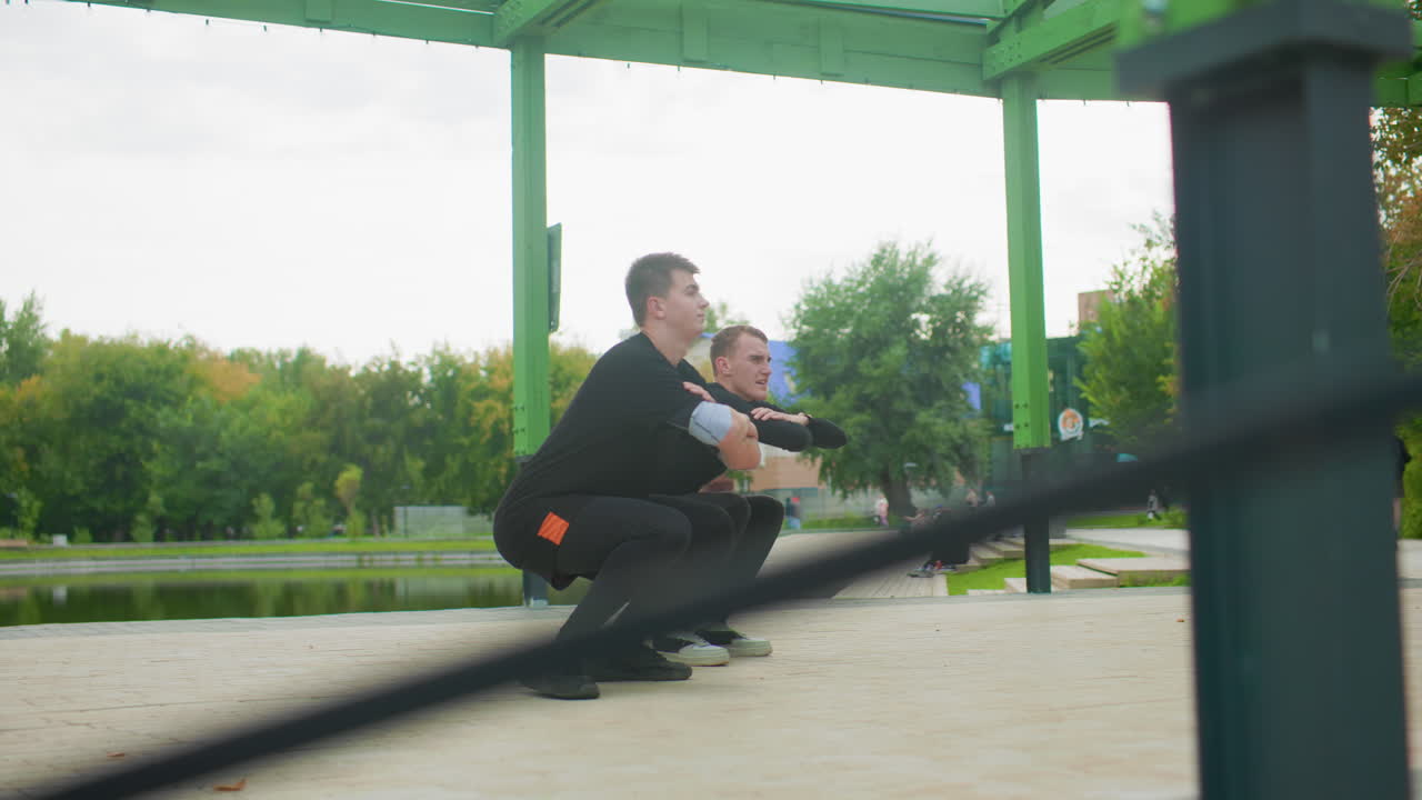 Istruttore in tenuta bianca che guida la tecnica dello squat in un padiglione, istruzioni di supporto chiare, modifica della postura, feedback del partner, dimostrazione dell'allineamento dei piedi, parco con vista lago, abbigliamento casual nero, atmosfera pomeridiana