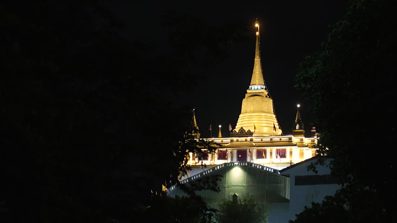 木々に囲まれた夜の黄金の仏塔、バンコクのワット・サケート（ゴールデンマウント寺院）の夜景、遠くに見える夜の仏教寺院。