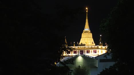 Golden-Pagoda-at-Night-surrounded-by-Trees,-The-Golden-Mount-Temple-in-Bangkok-at-Night,-Buddhist-Temple-at-Night-in-the-Distance