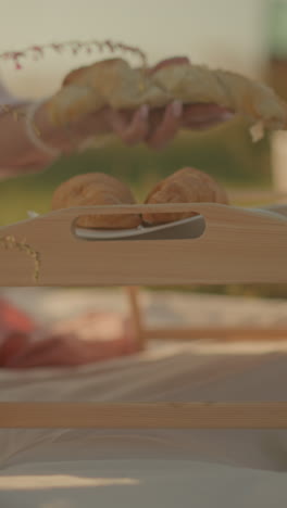 vista parcial de una mujer sentada en una alfombra de picnic al aire libre recogiendo croissants frescos de una bolsa de papel para colocarlos en platos encima de una mesa de picnic de madera, rodeada de una canasta tejida llena de flores