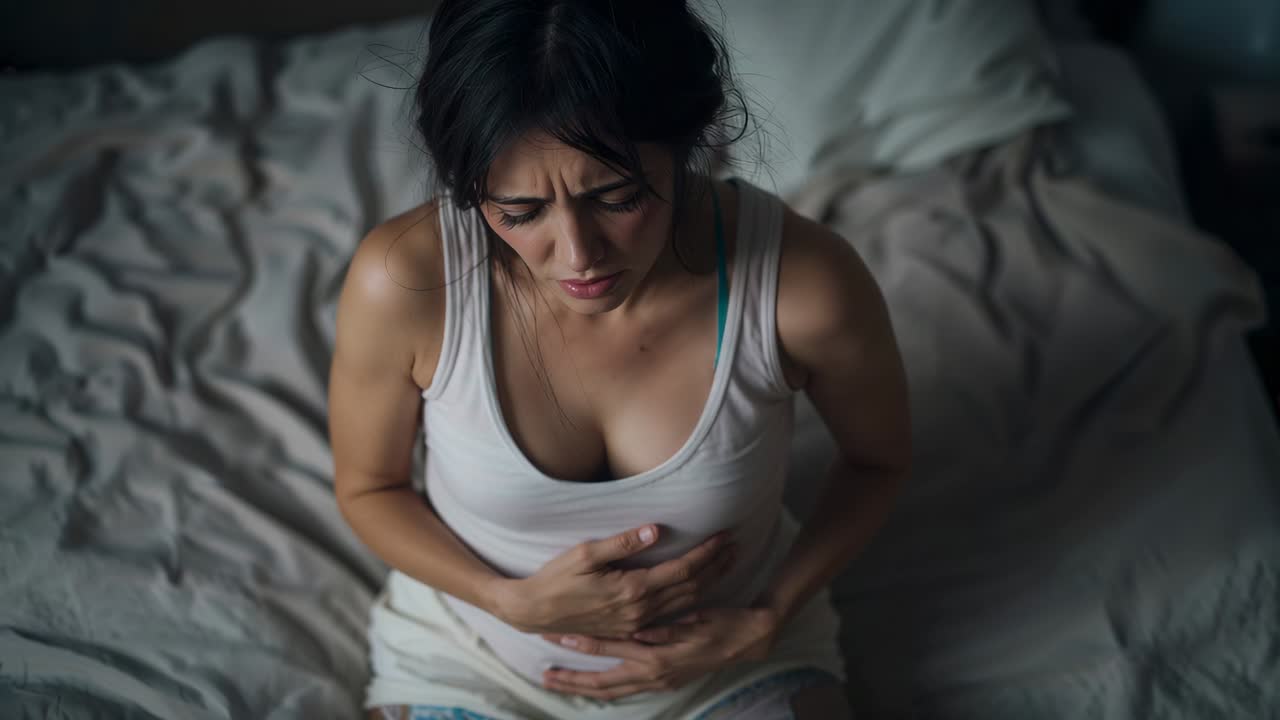 Holding woman grasping abdomen, pain starting at home on rumpled bed, tank top, bra strap visible