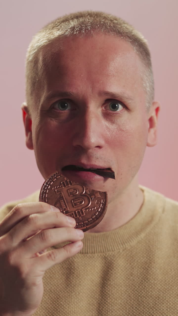 Man with Burr haircut bites piece of chocolate cookie with symbol of bitcoin looking in camera. Portrait of serious guy on blurred pink background closeup
