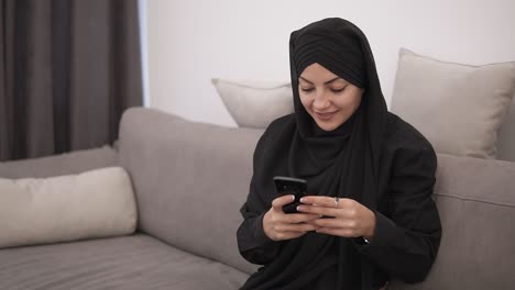 Schöne-Muslimische-Frau-In-Schwarzem-Hijab,-Die-Auf-Dem-Sofa-Sitzt-Und-Auf-Ihr-Smartphone-Schaut-Und-Lächelt