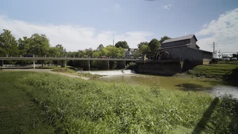 Cars-driving-over-bridge-next-to-Water-Mill-with-small-waterfall,-Jasper-Indiana