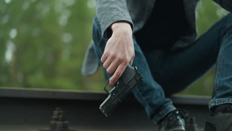 una vista cercana de una persona con una chaqueta gris, pantalones vaqueros azules y zapatos de lona negros sentados en una vía ferroviaria, sosteniendo una pistola con un agarre firme, el dedo en el gatillo y su cabeza en su otra mano
