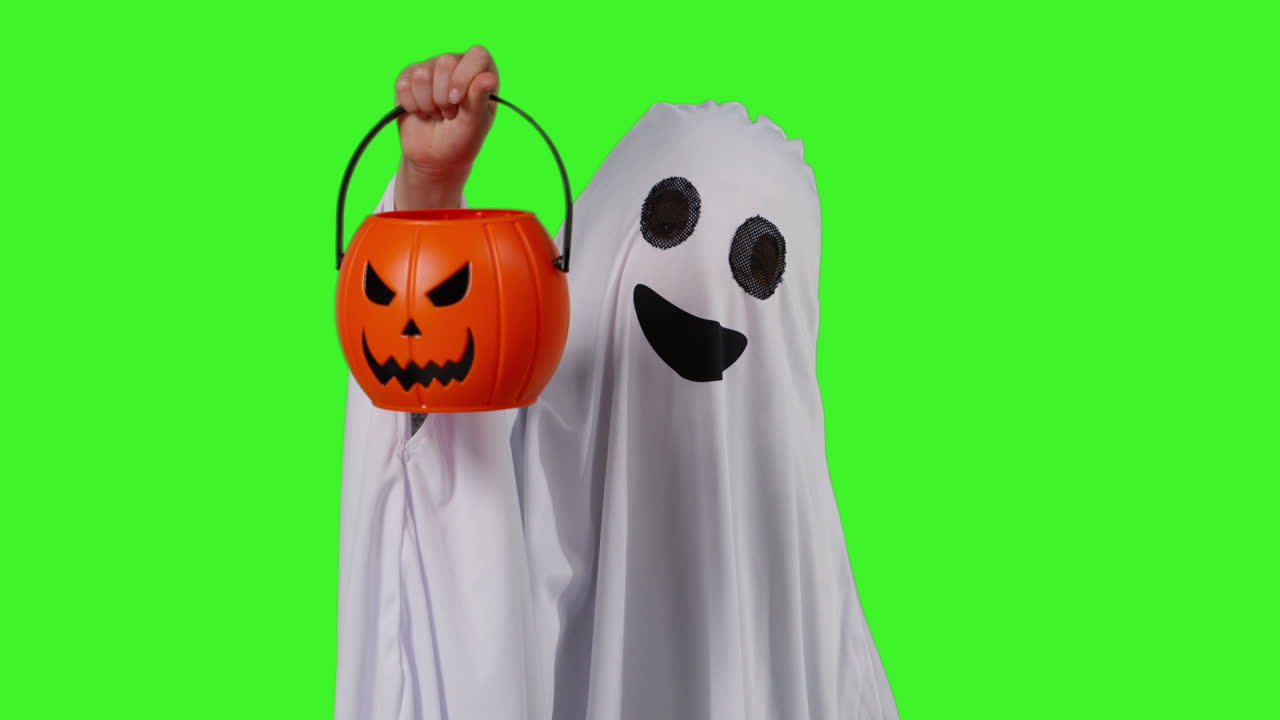 유령 의상을 입은 아이가 할로윈에 속임수를 치거나 할로윈에서 스튜디오 녹색 화면에 대항하여 호박 모양의 잭 오 랜턴 버을 들고 있습니다.