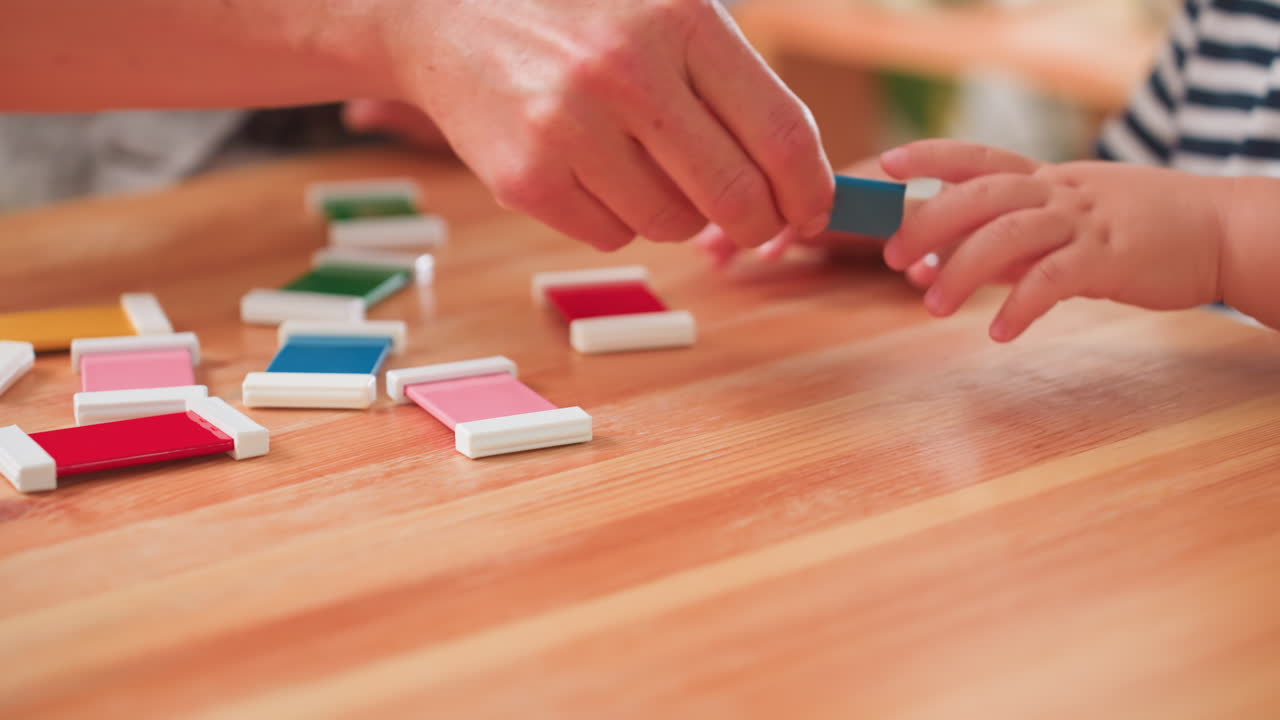 Adult helps kid arrange colorful block shapes by matching according to color on wooden table, guiding small hands, striped shirt visible, supportive learning moment fostering focus