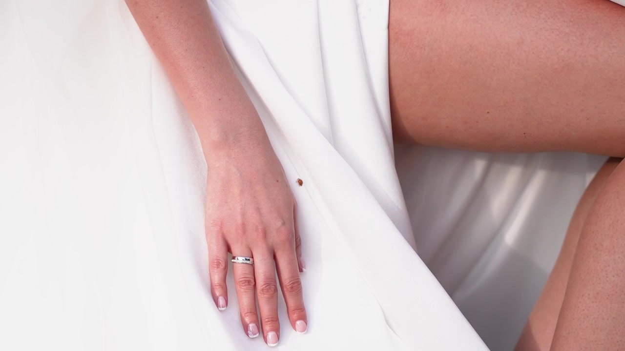 Primo piano dettagliato della mano di una donna che poggia su un tessuto bianco liscio, mostrando un anello elegante e un leggero movimento. L'illuminazione soffusa evidenzia la texture della pelle e una composizione delicata