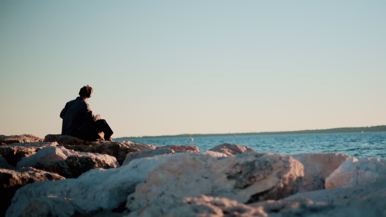 A man sitting alone on large rocks by the sea, looking toward the horizon under a clear sky