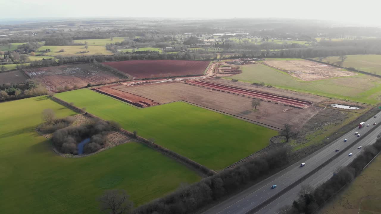 Drone footage showing construction work near motorway in Hampshire, England, with vehicles on road, red soil excavation areas, and surrounding green farmland under bright overcast daylight
