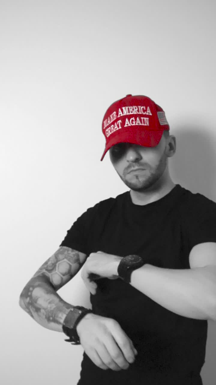 Grayscale footage of a man with tattoos putting on a red colored MAGA Make America Great again hat in slow motion, crossing his arms and looking driectly at the camera while nodding, man is confident.