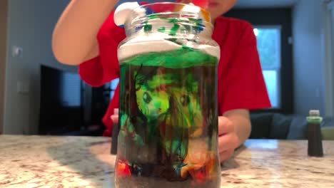 making an indoor rain cloud using shaving cream, food coloring, water, and a jar. young primary school aged boy demonstrating the fun, at home science experiment. 4k
