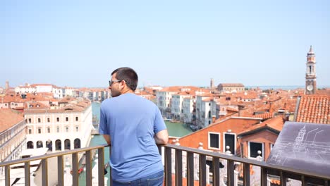 Rear-view-of-young-man-looking-at-beauty-of-Venice-cityscape-from-rooftop-of-Fondaco-dei-Tedeschi,-Italy
