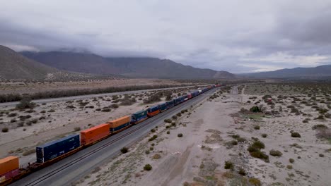 Imágenes-Aéreas-De-Drones-Del-Tren-De-Carga-En-El-Desierto-De-Palm-Springs-Con-Parques-Eólicos-En-El-Fondo,-Tiro-Lento-Hacia-Adelante