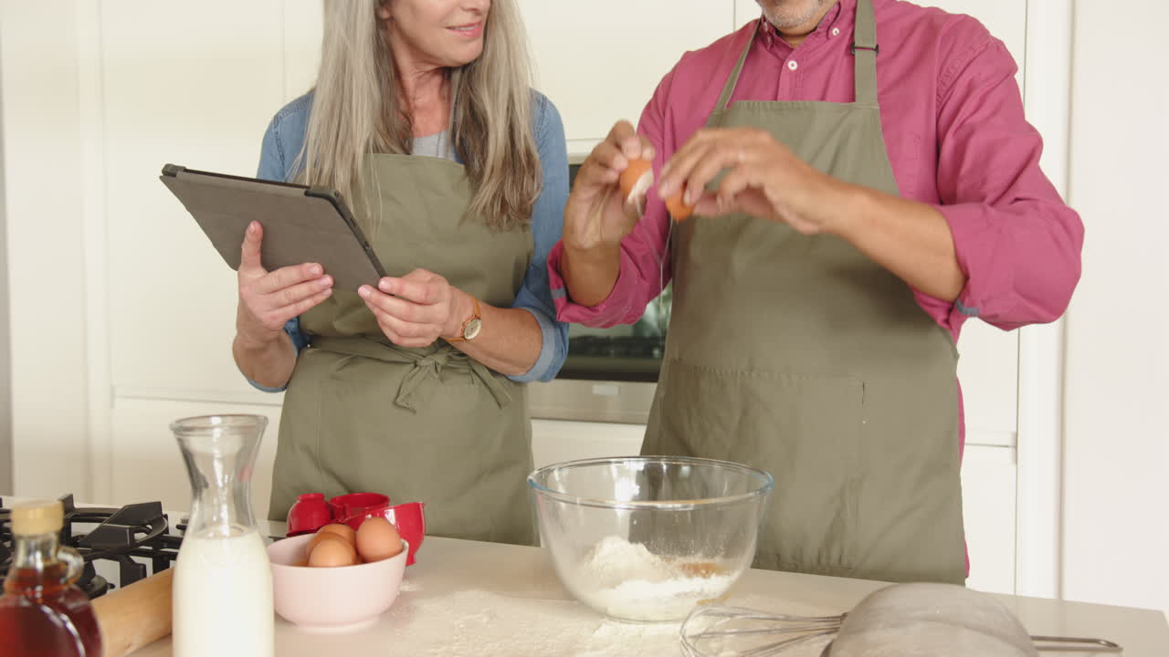 Baking together, multiracial senior couple using tablet and cracking eggs into mixing bowl, at home