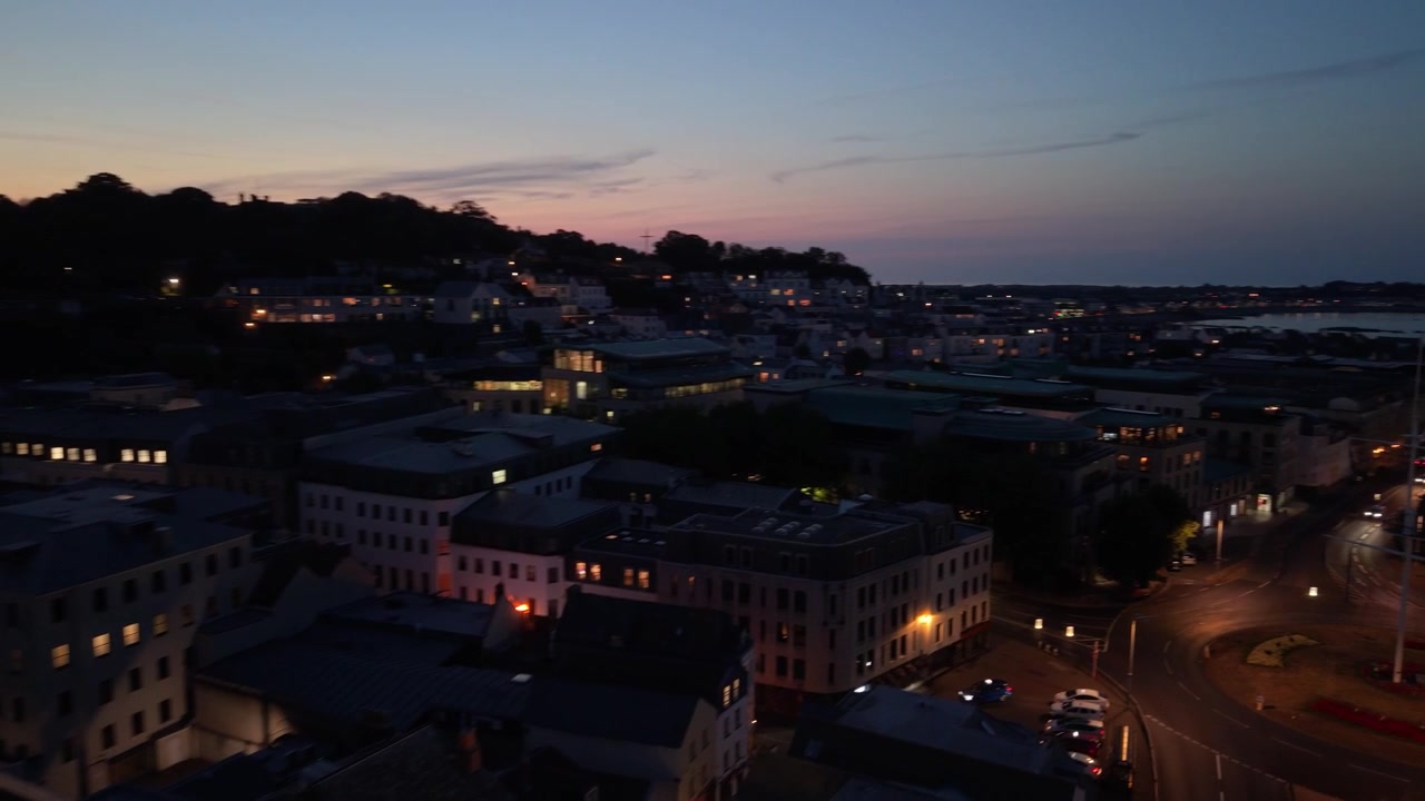 St Peter Port, Guernsey, Isole del Canale. Volo sopra il lungomare e gli edifici della città al crepuscolo con luci scintillanti contro il tramonto, con una panoramica per ammirare l'Esplanade di Glategny e la Baia di Belle Greve