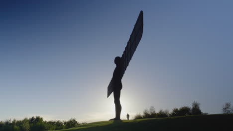 Angel-of-the-North-against-blue-sky,-silhouetted-man-with-dog-walks-along-the-path