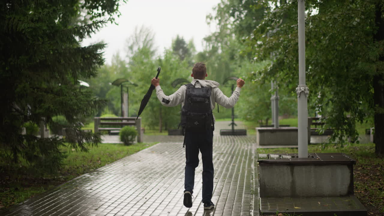 남자, 백팩, 젖은 공원 길을 걷고 있음, 우산을 아래로 잡고 있음, 반사되는 포장 도로에 빗방울이 맺힘, 빽빽한 녹색 나무와 가로등이 산책로를 둘러쌈, 느리고 외로운 발걸음, 흐린 날씨, 영화 같은 분위기