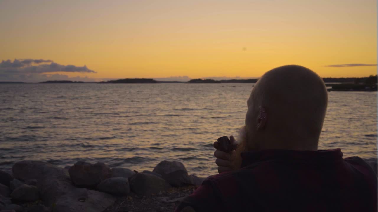 빈티지 파이프를 들고 담배를 피우는 어른 독성 구름 바람에 따라 움직이는 바다  ⁇ 의 다채로운 일몰을 바라보는 파도가 천천히 움직이는 하늘 몇 개의 구름과 함께 영화적인 평온의 느낌