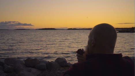hombre adulto sosteniendo una pipa vintage fumando nubes de toxinas moviéndose con el viento mirando la colorida puesta de sol junto al océano olas moviéndose en slowmo cielo con pocas nubes sensación cinematográfica de calma