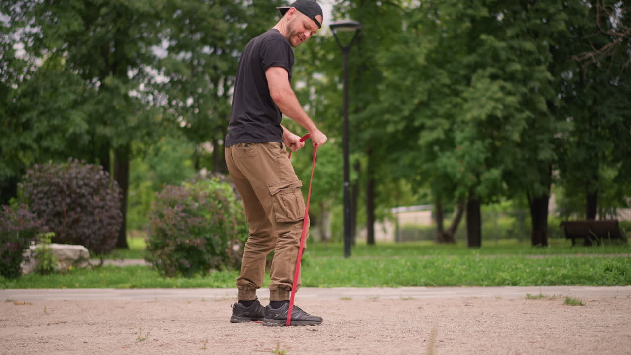 Uomo che fa stretching all'aperto, individuo di sesso maschile ancorato a un elastico di resistenza che si prepara per l'esercizio fisico all'aperto, uomo in abbigliamento casual che esegue esercizi di mobilità con un elastico di resistenza su un sentiero di ghiaia