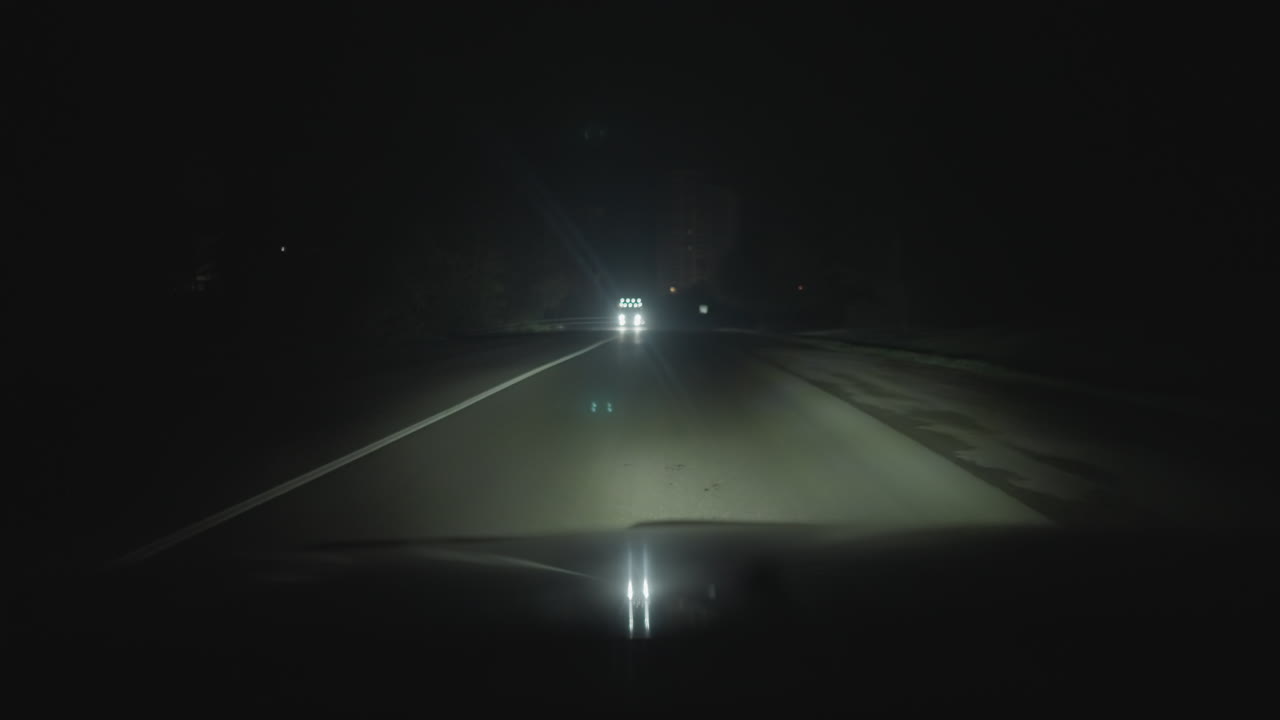 어두운 고속도로를 따라 다가오는 전조등, 밝은 빛이 강렬한 눈부심을 만들고 보닛에 반사돼요. 마주 오는 차량이 거리를 좁히고, 운전자는 속도를 줄이고, 길가의 표지판은 그림자 속으로 사라져요.