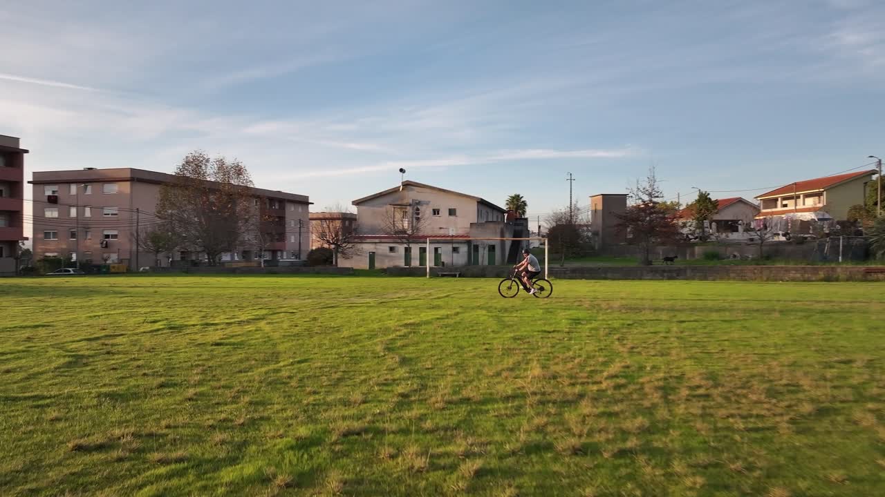 un ciclista cabalga a través de un campo de hierba de derecha a izquierda, con un disparo panorámico que revela un poste de la portería al atardecer.