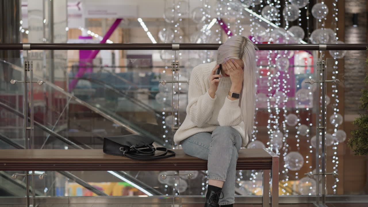 urban dweller seated on bench in mall walkway, holding phone to ear with eyes closed in exhaustion, wearing sweater, jeans and boots, black purse beside her against glass railing and festive lights