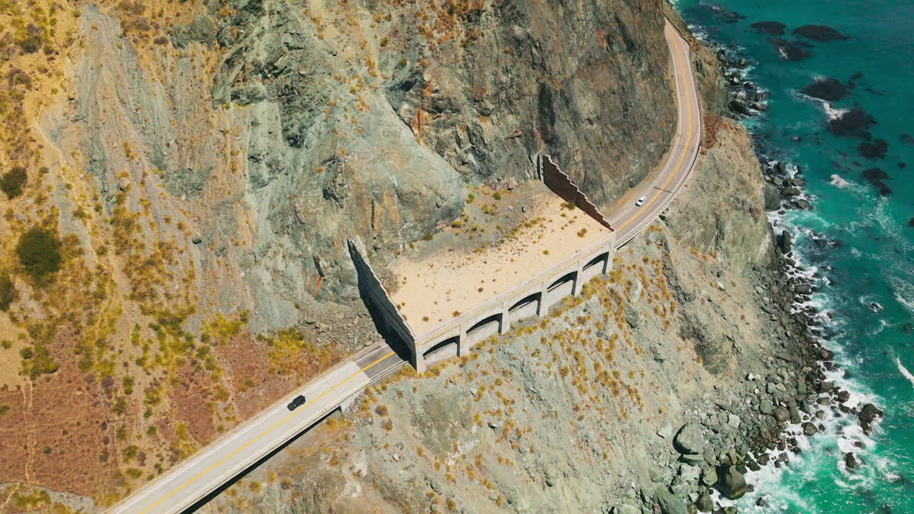 Cars moving by the dangerous road in the mountains of pacific shore. Turquoise ocean waves crush by the rocks. Top view.