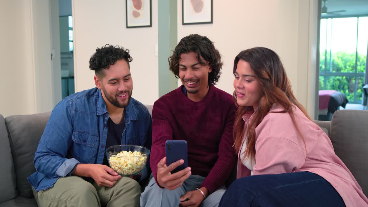 Amigos diversos se acomodando no sofá cinza, pegando o celular, tocando e rolando um clipe engraçado e pegando pipoca.