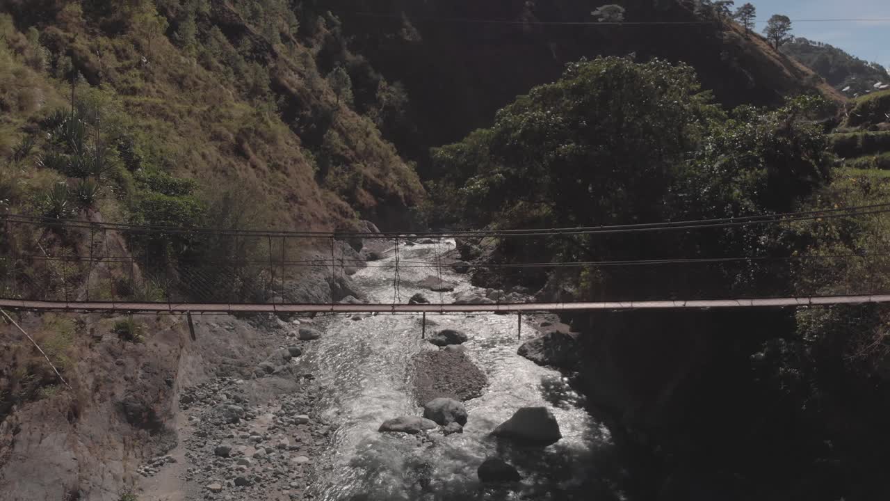Metal wire suspension bridge spanning over mountain water rocky river flowing away connecting trail trees grass mountain nature aerial wide fixed position Benguet Philippines