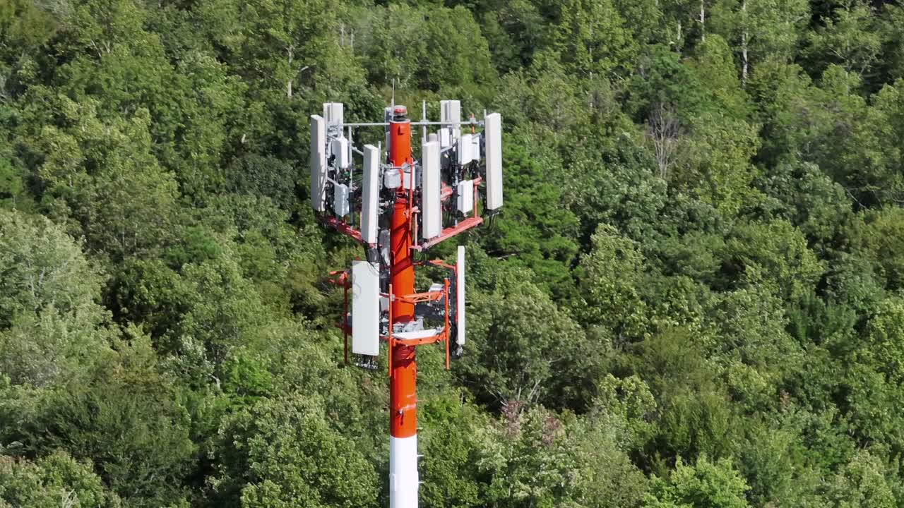 Red and white telecommunication tower in forest landscape of American town. Aerial close up orbiting shot. Sunny summer day in hills woodlands of Virginia