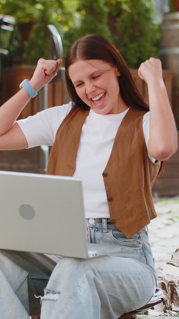 노트북에서 일하는 행복한 젊은 여성이 즐거움으로 비명을 지르며 도시 거리에서 손을 들어 올립니다.