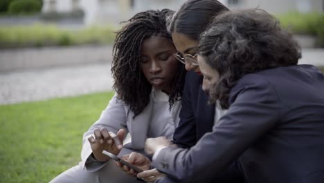 mujeres jóvenes serias sentadas en el prado y mirando el teléfono inteligente