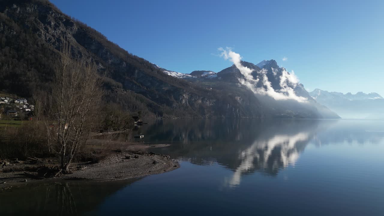 Drone clip moving towards misty snow topped mountain range, on edge of calm lake, on bright sunny day