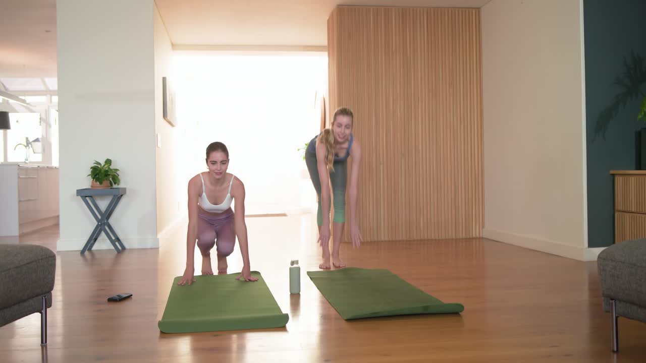 Amigas entrando em casa carregando colchonetes de yoga, colocando o celular e a garrafa, ajoelhando-se e começando a fazer yoga