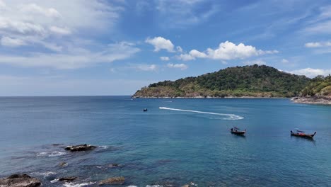 drone footage captures longtail boats navigating clear waters along phuket's rocky coastline under bright, sunny skies