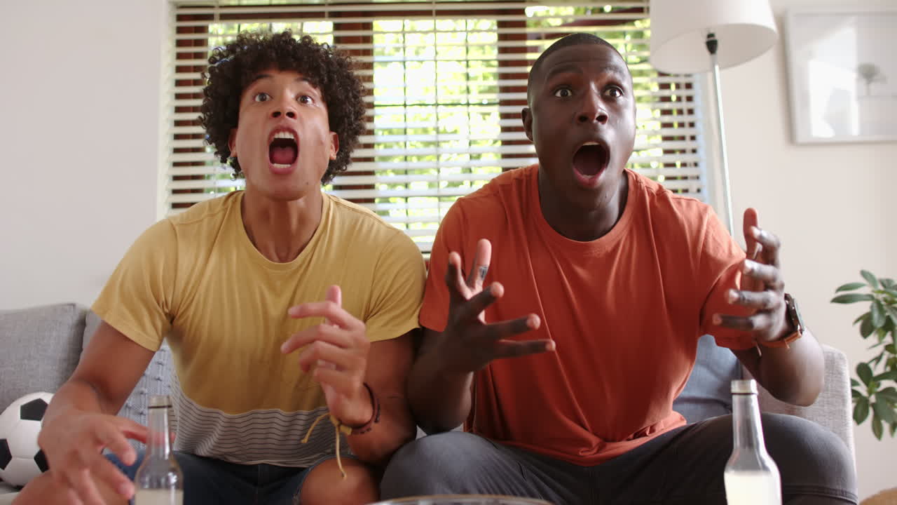 Watching sports game, two multiracial male friends reacting with excitement and frustration, at home