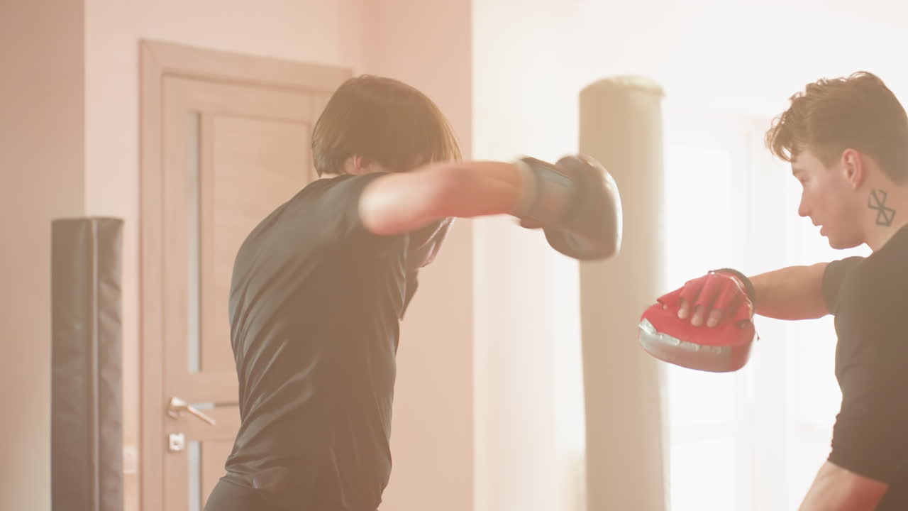 Boxer in black shirt wearing gloves throws strong punch during intense training session, showing athletic discipline, focus, power, and strength in combat practice inside gym with martial arts energy